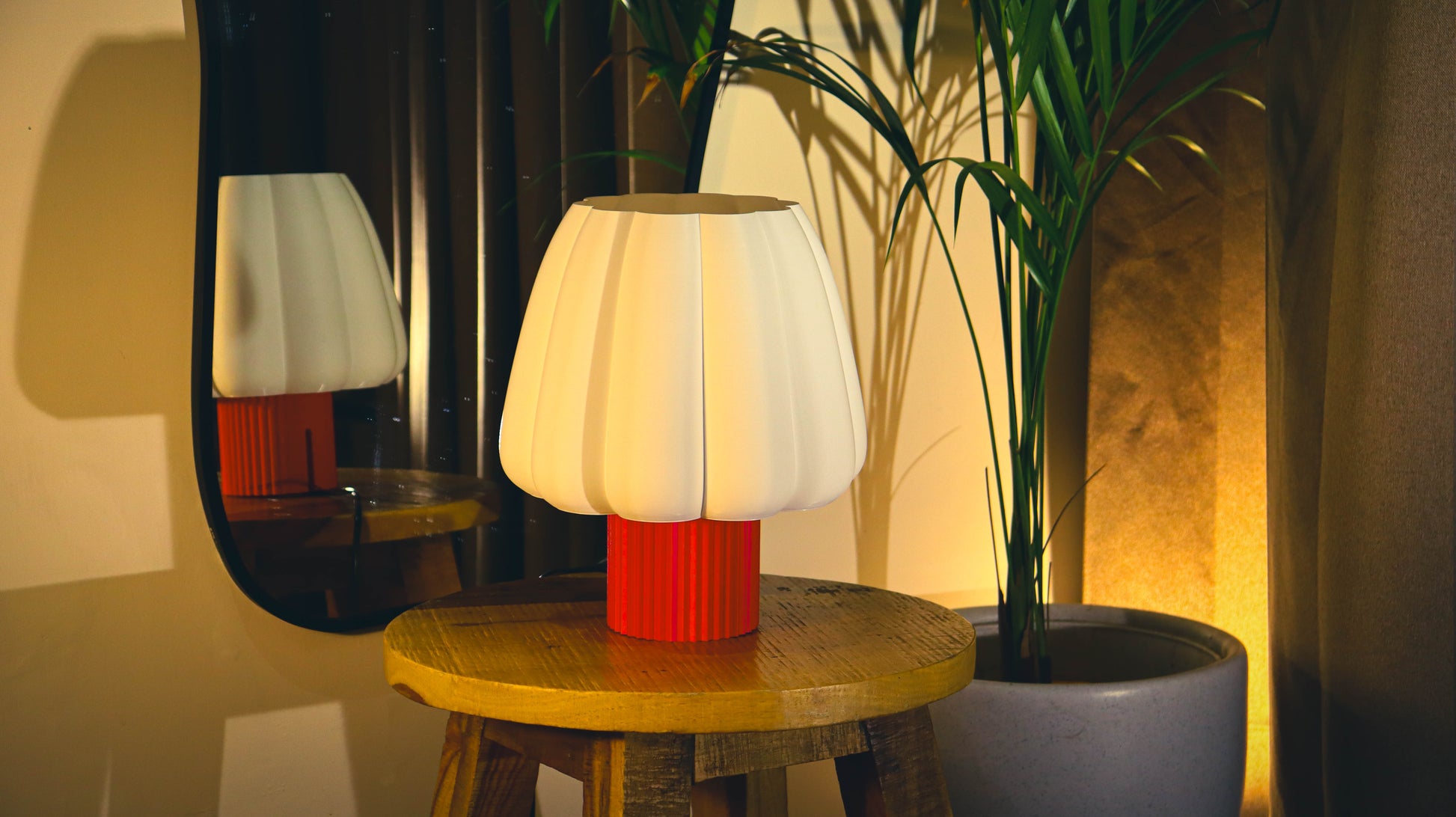 Table lamp with a red base and white shade on a wooden table, next to a plant and mirror.