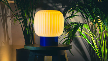 Cozy warm-lit table lamp, ribbed white shade, blue base, tropical plant backdrop