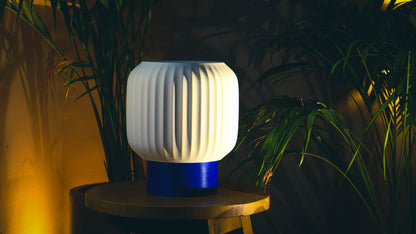 White textured lamp with blue base on a wooden stool in a dark setting with plants